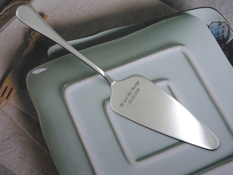 Puede incluir: Un servidor de pastel plateado con un mango largo y una hoja ancha y en &aacute;ngulo. El servidor est&aacute; grabado con el texto "Mr and Mrs Martyn 25.05.2019". Descansa sobre una pila de platos cuadrados verde claro.