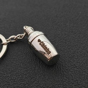 Pode incluir: Um porta-chaves prateado em forma de shaker de cocktail, com o nome "Chinba" gravado na lateral. Um símbolo de coração precede o nome. O shaker tem um design em espiral no topo e está preso a uma corrente e porta-chaves prateados.