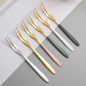 Pode incluir: Seis garfos de sobremesa com um design moderno. Os garfos têm dentes dourados e cabos com uma variedade de cores, incluindo preto, branco, rosa e verde. Os garfos estão dispostos diagonalmente sobre uma superfície cinza e branca.