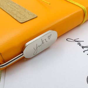 May include: A close-up of a silver bookmark with the name "Yuki" engraved on it, with a heart symbol next to the name. The bookmark is attached to a bright orange leather notebook.