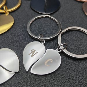 Op de afbeelding: Close-up van zilveren hartvormige sleutelhangers, in twee helften gesplitst, elk met een letter gegraveerd. Eén sleutelhanger toont de letters "N" en "C". Andere sleutelhangers in goud en zwart zijn ook zichtbaar. De sleutelhangers liggen op een donkere ondergrond.