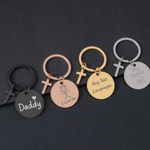 May include: A selection of keyrings with circular pendants and cross charms. The keyrings are in black, rose gold, and gold colours. Each pendant has a different inscription, including "Daddy", "I Love You", and "My Family".