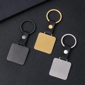 May include: Three square keychains in gold, silver, and gunmetal. Each keychain has a square metal plate attached to a black leather strap with a metal ring. The keychains are on a black surface.