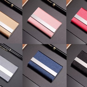 以下が含まれることがあります： ゴールド、ピンク、レッド、グレー、ブルー、ブラックなど、様々な色の名刺入れのコレクション。 各ホルダーは長方形で、中央にシルバーのストライプが入っています。 金属製と思われる名刺入れが並んでいます。