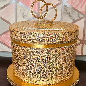 May include: Gold wedding card box with a decorative ring topper. The box is covered in iridescent sequins and has a gold ribbon around the middle.