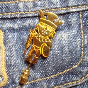 May include: Gold-colored pin featuring a stylized figure of a person with a headdress. The figure is wearing a necklace and holding a shield. The pin is attached to a denim jacket.