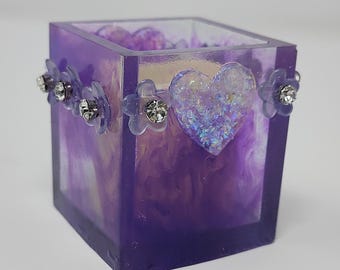 Puede incluir: Un portavelas cuadrado de resina morada con adornos de corazón y flores. El portavelas tiene un efecto veteado con tonos morados y blancos. El corazón está relleno de purpurina iridiscente. Las flores tienen pétalos transparentes y centros de pedrería.