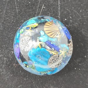 May include: A clear, round resin paperweight with embedded seashells, starfish, and sand dollars in shades of blue, turquoise, and gold. The paperweight has a glossy finish and a nautical theme.