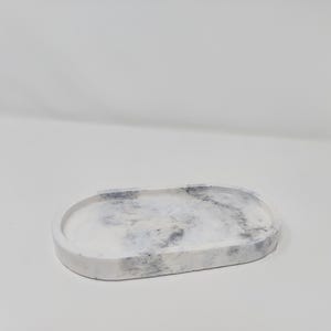 May include: Oval-shaped decorative tray with a marbled design. The tray is primarily white with gray and black veining, creating a natural stone appearance. The edges are smooth and rounded, and the tray is made of a solid material.