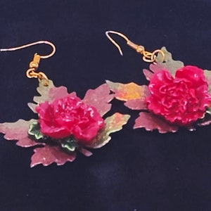 May include: A pair of earrings featuring a red flower with green leaves. The earrings are made of metal and have a gold finish.