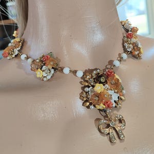 Puede incluir: Un collar de estilo vintage con racimos de flores coloridas en tonos naranja, amarillo y blanco, acentuado con perlas de imitación y pedrería. El collar incluye un colgante de lazo dorado con pedrería transparente y una cadena de cuentas blancas.