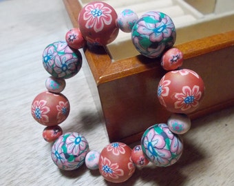 Pode incluir: Um bracelete feito de contas com padrão floral vermelho, rosa e verde. As contas são enfiadas em um cordão elástico transparente.