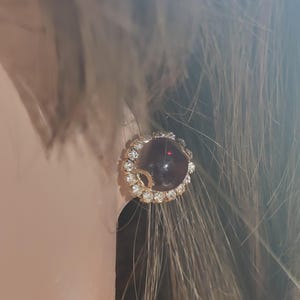 May include: A pair of gold earrings with a round, dark red stone surrounded by clear rhinestones.