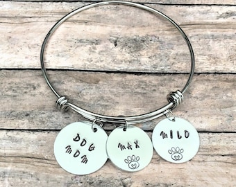 May include: A silver-colored bangle bracelet with three round charms. The charms have stamped text that reads "DOG MOM", "MAX" with a paw print, and "MILO" with a paw print. The bracelet is on a wooden surface.