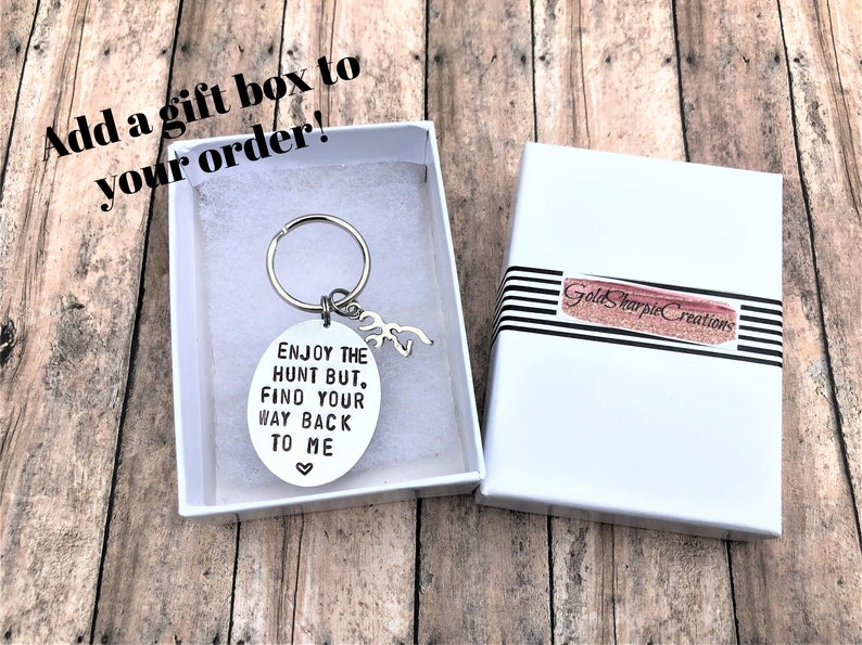 May include: A silver key chain with a round charm that says "Enjoy the hunt but, find your way back to me" with a small heart symbol. The key chain is in a white gift box with a black and white striped ribbon.