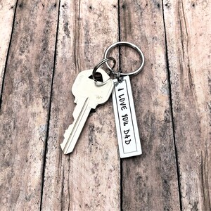 May include: Silver key ring with a silver key and a silver tag that says "I Love You Dad". The keyring is on a wooden surface.