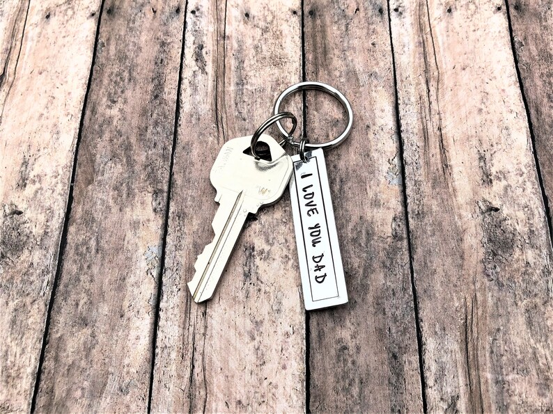 May include: Silver key ring with a silver key and a silver tag that says "I Love You Dad". The keyring is on a wooden surface.