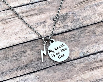 May include: Silver-toned necklace with a chain and two charms. One charm is a lightning bolt, and the other is a circular pendant with the phrase "My heart is on the line" in black lettering. The necklace is displayed on a wooden surface.