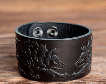 Puede incluir: Pulsera de cuero negro con un diseño en relieve de dos lobos enfrentados. La pulsera tiene un cierre de presión.