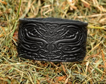 Puede incluir: Pulsera de cuero negro con un intrincado diseño de nudo celta. El diseño presenta dos dragones entrelazados con sus cabezas enfrentadas.