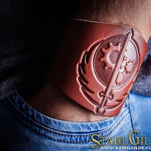 Puede incluir: Una pulsera de cuero marrón con un diseño estilizado que presenta una espada, engranajes y un símbolo alado. El diseño recuerda a la franquicia de videojuegos Fallout.