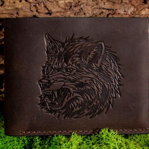 Könnte beinhalten: Eine dunkelbraune Ledergeldbörse mit einem geprägten Wolfskopf-Design. Die Geldbörse hat eine rechteckige Form mit genähten Kanten. Der Wolfskopf ist aufwendig detailliert, mit scharfen Zähnen und einem wilden Ausdruck.