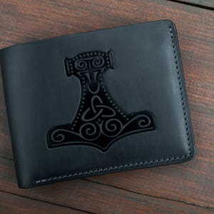 Puede incluir: Cartera de cuero negro con una imagen en relieve detallada del martillo de Thor, Mjolnir. La cartera es rectangular y cerrada, con costuras visibles en los bordes. Fondo de madera oscura.