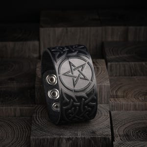 May include: Black leather cuff bracelet featuring a silver pentagram design within a circle. The bracelet has an embossed Celtic knot pattern and three silver snap closures. The cuff is displayed on a dark wooden surface.