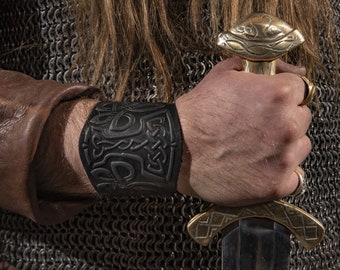 Puede incluir: Un brazalete de cuero negro con un diseño de nudo celta. El brazalete se lleva en la muñeca de una persona que sostiene una espada.