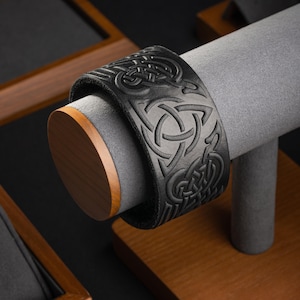 Puede incluir: Un brazalete de cuero negro con un diseño de nudo celta. El brazalete se muestra en un soporte de madera.