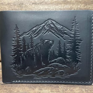May include: Black leather wallet featuring a mountain scene with a bear, trees, and rocks. The design is embossed on the front of the bi-fold wallet. The wallet has a stitched border.