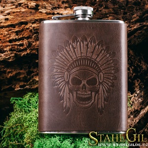 May include: A brown leather-covered flask with a silver top and a skull design with a Native American headdress. The flask has the text "Stahl Gilde" and "www.stahlgilde.com" on the bottom.