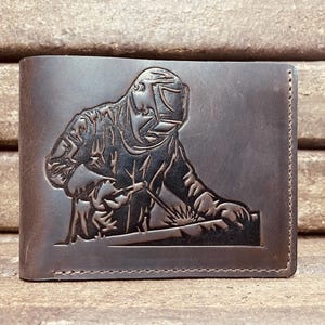 May include: A brown leather wallet with a detailed embossed image of a welder at work. The wallet is rectangular and features a bi-fold design. The image shows the welder with a welding mask and tools.