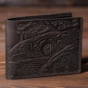 Puede incluir: Una cartera bifold de cuero marrón oscuro con una escena de paisaje en relieve que muestra una puerta redonda en una colina cubierta de hierba, una chimenea con humo, árboles y un camino de piedra, con costuras visibles en los bordes.