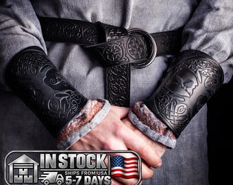 May include: A pair of black leather bracers featuring embossed Celtic knotwork patterns and a matching black leather belt with a large metal ring buckle, worn over a light gray textured long-sleeved garment with rolled cuffs.