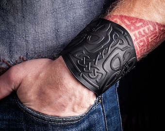 Puede incluir: Un brazalete de cuero negro con un intrincado diseño de nudo celta. El brazalete se lleva en la muñeca.