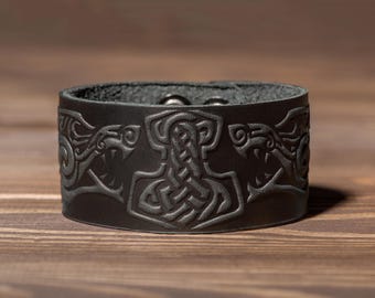 Puede incluir: Una pulsera de cuero negro con un diseño en relieve detallado. El diseño presenta un martillo de Thor central flanqueado por dos cabezas de lobo estilizadas. La pulsera se muestra sobre una superficie de madera.