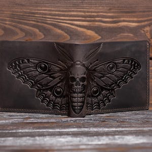 Op de afbeelding: Een donkerbruine lederen bifold portemonnee met een gedetailleerd reliëfontwerp van een mot met een schedelpatroon op het lichaam. De portemonnee heeft zichtbare stiksels langs de randen en een gladde, matte afwerking met ingewikkelde vleugeldetails.