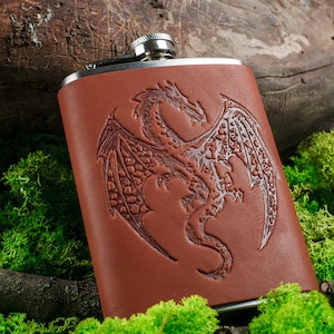 Puede incluir: Una petaca de cuero marrón con tapa plateada y un diseño de dragón grabado en relieve.
