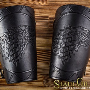 Puede incluir: Un par de protectores de brazo de cuero negro con un diseño en relieve de una cabeza de lobo feroz con una corona de espinas. Los protectores de brazo se aseguran con hebillas y correas.