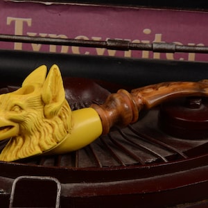 May include: A yellow and brown wooden pipe with a wolf head design. The pipe is sitting on a brown surface.