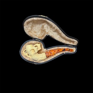 May include: A white and orange carved pipe with a detailed design, resting in a black velvet lined case.