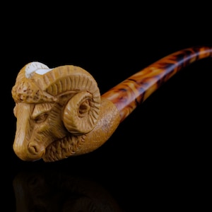 May include: A wooden pipe with a ram's head carved into the bowl. The pipe is brown and has a mottled finish. The ram's head is detailed with horns, eyes, and a nose.