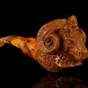May include: A brown wooden pipe carved in the shape of a ram's head. The pipe has a long, curved stem and a bowl shaped like the ram's head. The ram's horns are curled and detailed.