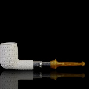 May include: A white ceramic pipe with a textured bowl and a brown, amber-colored stem. The pipe is lying on a black surface.