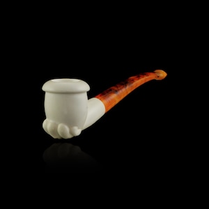 May include: A white ceramic pipe with a brown and orange stem. The bowl of the pipe is shaped like a hand.