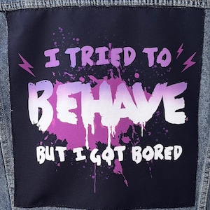 Peut inclure: Veste en jean bleu clair avec un grand patch noir dans le dos. Le patch affiche le texte "I TRIED TO BEHAVE BUT I GOT BORED" en blanc et violet, avec des éclairs.