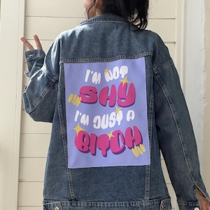 Può includere: Giacca di jeans blu chiaro. La parte posteriore della giacca presenta una toppa viola con la scritta "I'M NOT SHY I'M JUST A BITCH" in lettere rosa e bianche. Sono presenti anche stelle gialle.