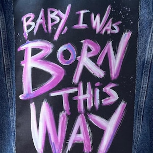 Op de afbeelding: Een spijkerjack met een zwarte patch die op de achterkant is genaaid. De patch heeft de woorden "Baby, I was born this way" erop geschreven in roze en witte verf.