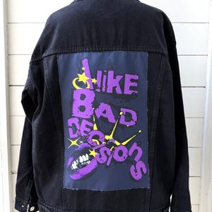 Könnte beinhalten: Eine schwarze Jeansjacke mit einem großen rechteckigen Aufnäher auf der Rückseite. Der Aufnäher zeigt den violetten Schriftzug "I LIKE BAD DECISIONS" mit gelben Sternen und einer Grafik aus violetten Lippen auf dunkelblauem Grund.